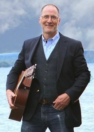 Chris Mahan standing by the lake, holding an acoustic guitar
